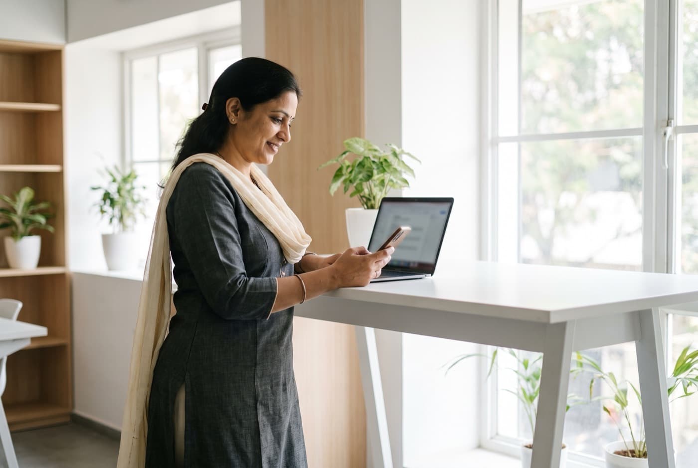 A business owner in a bright office checks her numbers on her laptop and phone.